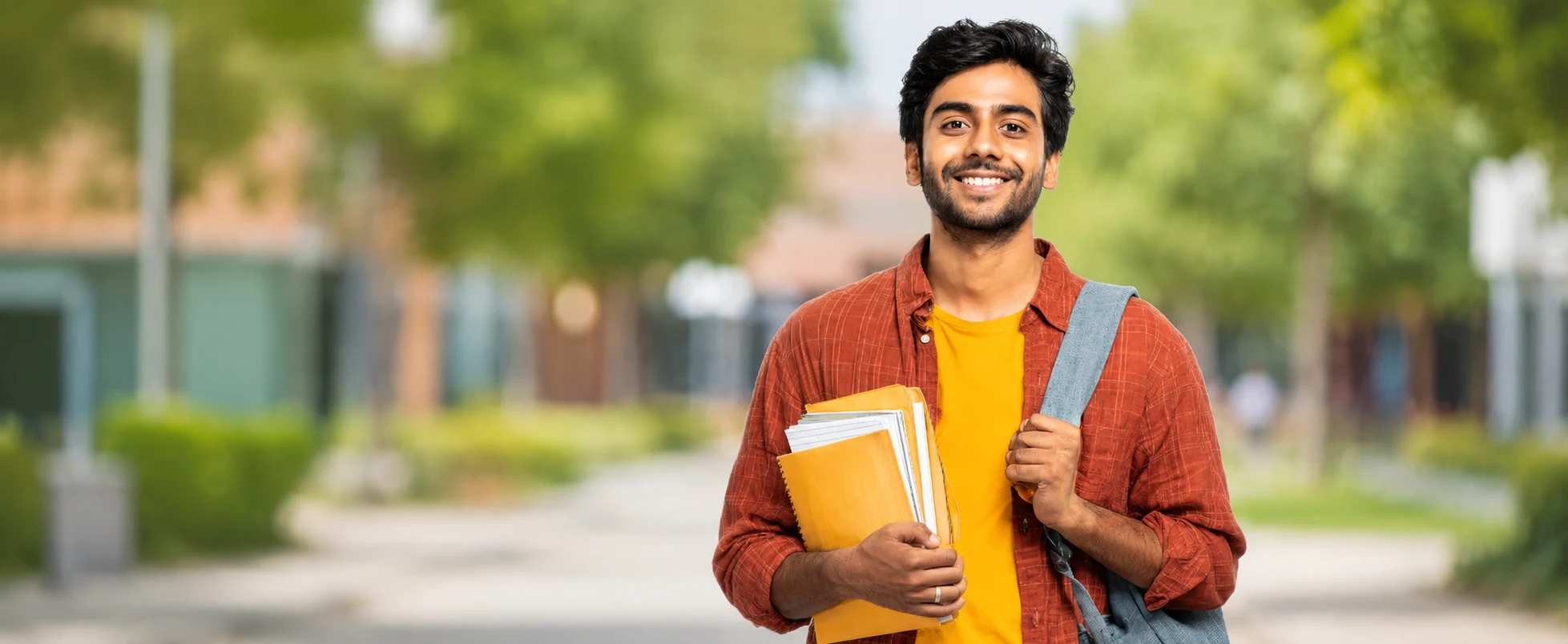 College-campus-banner Smiling Indian college boy holding books with blurred outdoor campus background